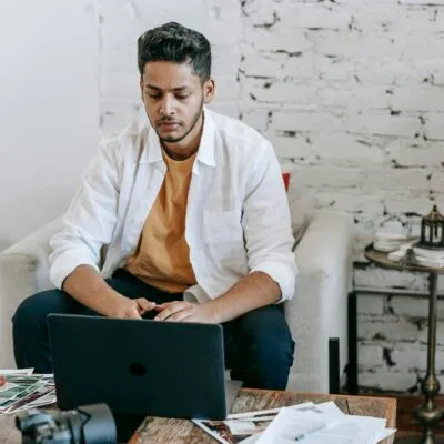 Creative young Hispanic male photographer sitting at table with photo camera and printed photos and working on laptop in modern loft style workplace at home