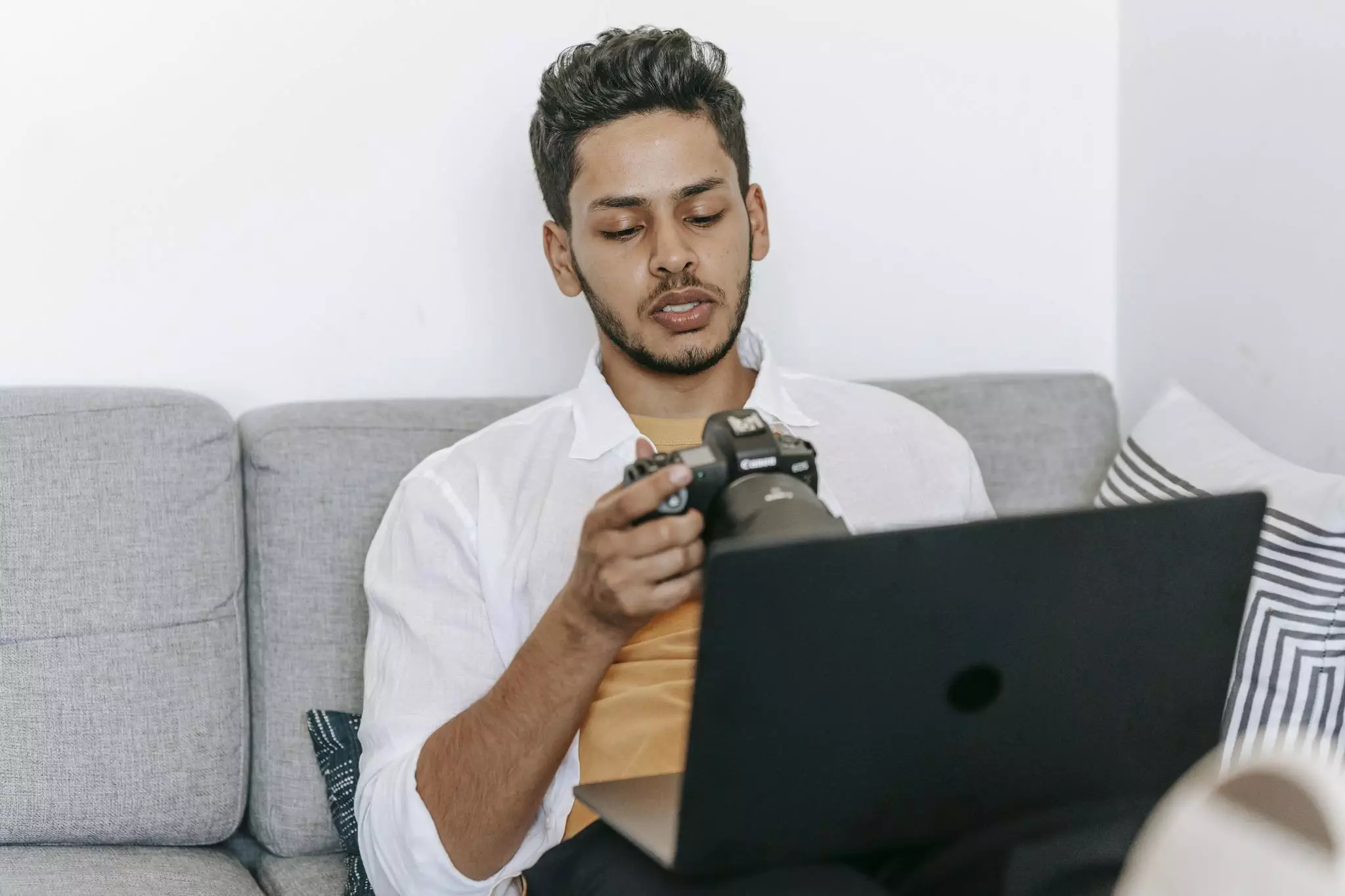 Young bearded ethnic male photographer checking pictures on photo camera while sitting with laptop on sofa and working on freelance project
