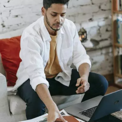 Young Hispanic male designer taking planner from table with laptop and pictures while working on project in modern loft style workplace