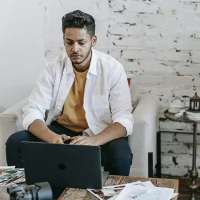 Creative young Hispanic male photographer sitting at table with photo camera and printed photos and working on laptop in modern loft style workplace at home